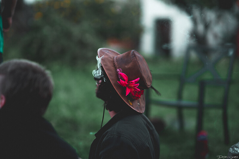A man wearing an earth brown hat with a vivid pink flower in it smoking in Berkeley, CA