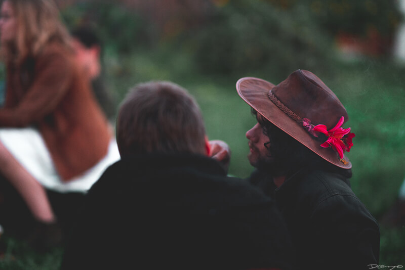 A man wearing a brown hat with a vibrant pink and purple flower in it seen smoking from behind in Berkeley, CA